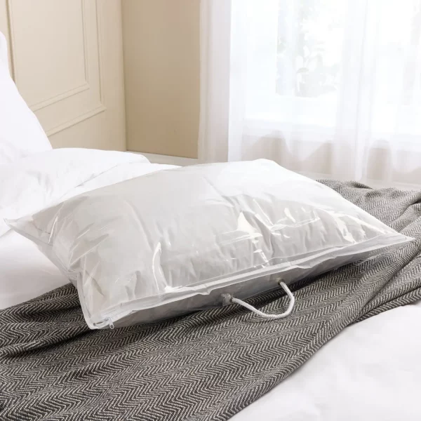 A transparent plastic storage bag containing a white pillow. It's sat on a grey blanket that's running across a bed. A window is visible in the background.