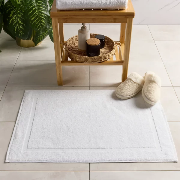 A Hotel Pure Luxury picture frame bath mat lying on a bathroom floor. There are also some fluffy slippers positioned in the right-hand corner. Visible at the back is a table with a basket at on the bottom shelf, in which sits a soap dispenser and a black pot. There are towels sitting on top of the table and a plant just about visible at the back of the room.