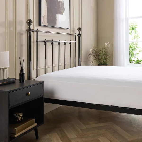 A white Hotel Pure Luxury cotton percale fitted sheet on a mattress that's on a metal framed bed. A dark wood cabinet with a lamp on top sits next to the bed. A painting is visible on the wall above the bed and you can just about see a window on the far-right hand side of the image.