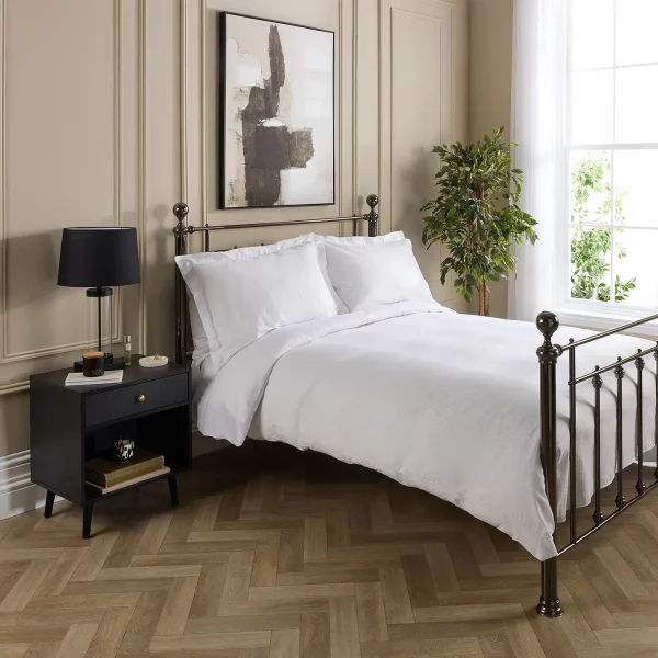 A Hotel Pure Luxury cotton rich percale satin stripe duvet cover on a bed, complete pillows and pillowcases. There is a dark wood bedside cabinet with a lamp sitting on top of it next to the bed. You can see a contemporary painting above the metal framed bed.