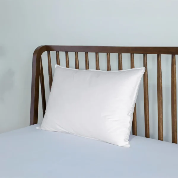 A white Martex Naturals duck feather and down pillow on a bed propped against a brown wooden headboard on a bed with a light-coloured fitted sheet covering the mattress.