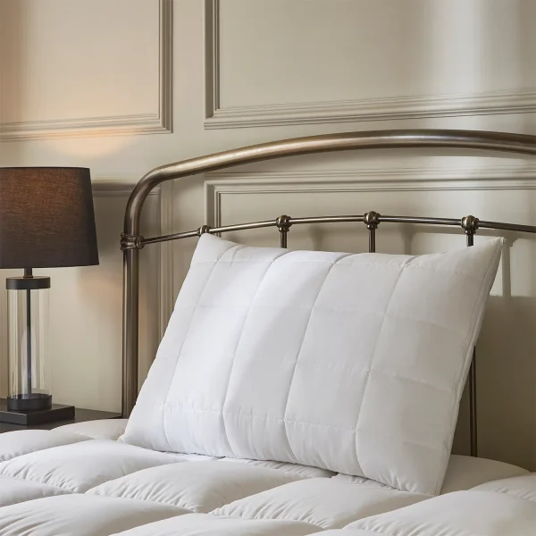 A Hotel Pure Luxury Premium Suite waterproof pillow protector covering a pillow. It's sitting on a bed with a metal frame headboard and a mattress topper. There is a lamp visible on the left-hand side.