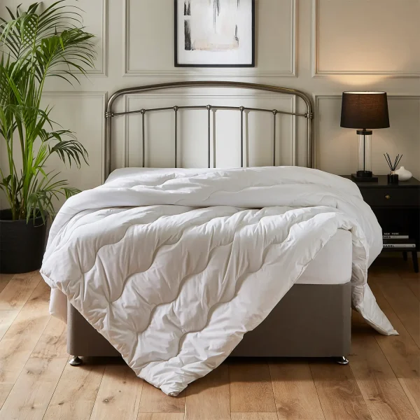 A Hotel Pure Luxury Premium Suite 10.5 tog duvet sitting on a bed with a metal frame headboard. It's in a modern bedroom with a lamp, plant and picture on the wall.