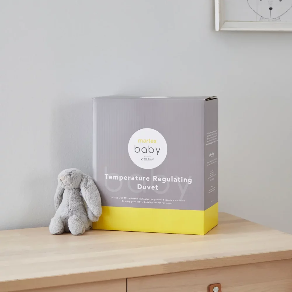 Grey and yellow packaging for a Martex Baby temperature regulating 4 tog duvet for use in a cot bed. The packaging is sitting on a chest of drawers and there is a small grey toy bunny sitting next to it.