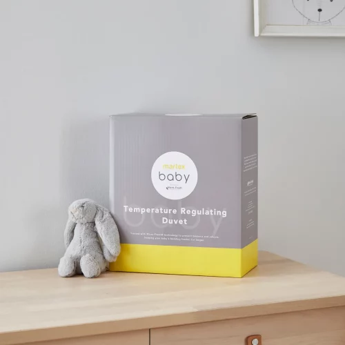 Grey and yellow packaging for a Martex Baby temperature regulating 4 tog duvet for use in a cot bed. The packaging is sitting on a chest of drawers and there is a small grey toy bunny sitting next to it.