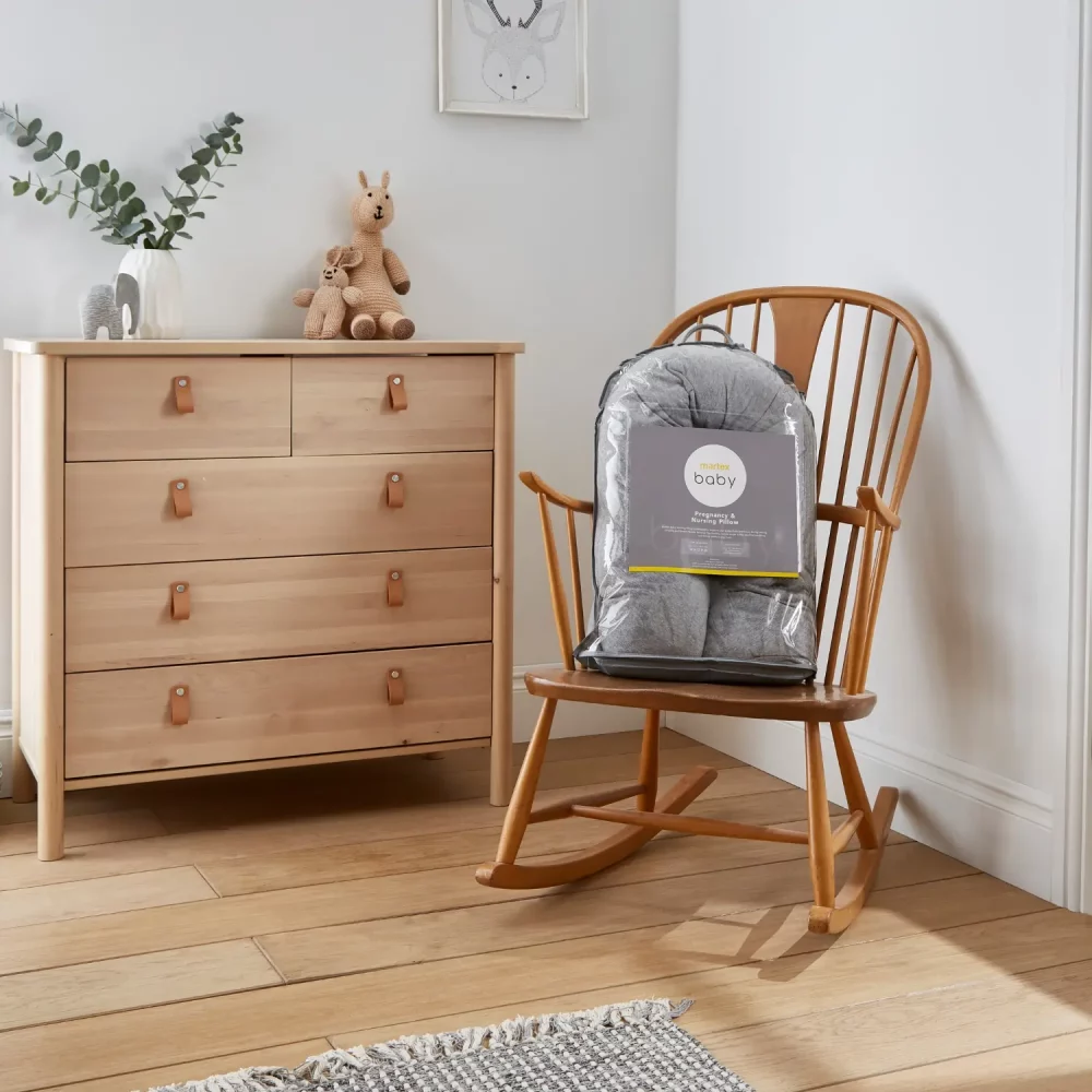 Martex Baby pregnancy and nursing pillow in grey. It's in its packaging and is sitting on a wooden chair in a room with a wooden floor and a wooden chest of drawers next to the chair.