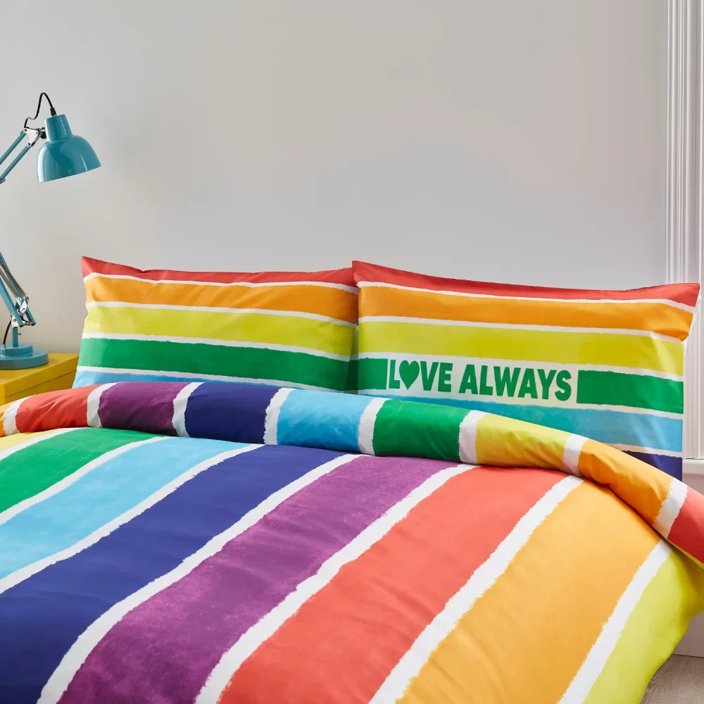 A multi-coloured rainbow Pride themed duvet cover and pillowcase set from Martex Pride covering a bed in a modern bedroom. It says 'Love Always' on one of the pillowcases.
