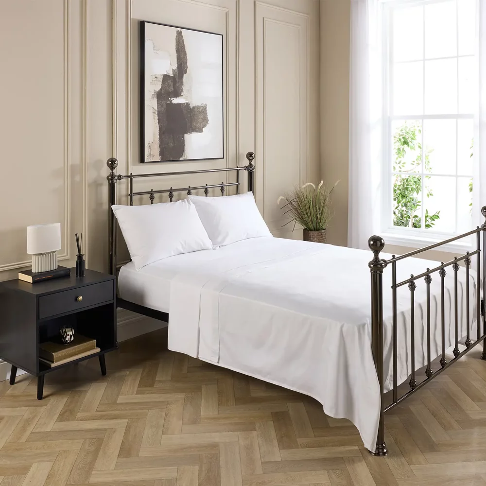 A Hotel Pure Luxury cotton rich percale flat sheet on a bed, complete with fitted sheet, pillows and pillowcases. There is a dark wood bedside cabinet with a lamp sitting on top of it next to the bed. You can see a contemporary painting above the metal framed bed.