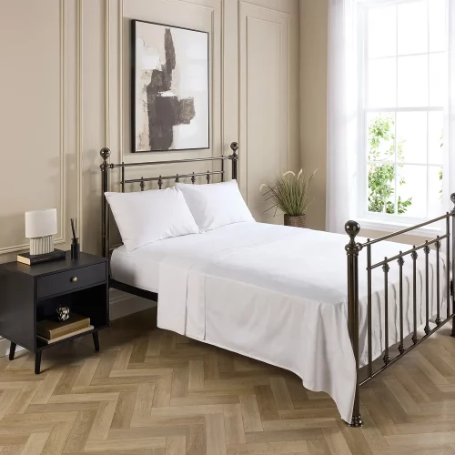 A Hotel Pure Luxury cotton rich percale flat sheet on a bed, complete with fitted sheet, pillows and pillowcases. There is a dark wood bedside cabinet with a lamp sitting on top of it next to the bed. You can see a contemporary painting above the metal framed bed.
