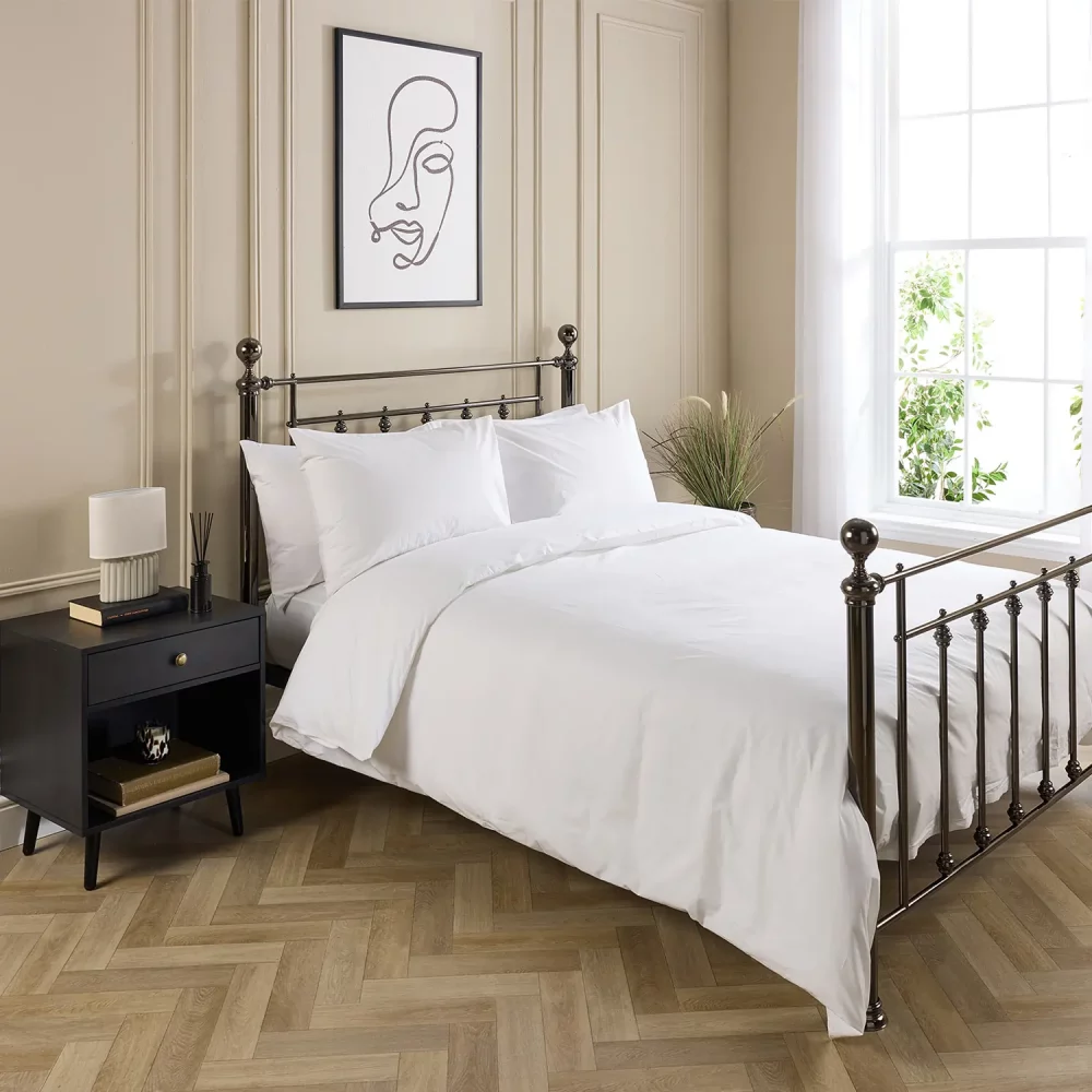Hotel Pure Luxury cotton rich percale extra long duvet cover on a metal framed bed in a contemporary bedroom. There are also pillows on the bed. There is a dark wood cabinet with a lamp on top on the left-hand side of the room, with a painting above the bed. There is a big window on the right-hand side of the room.