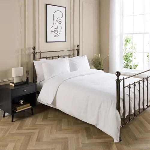Hotel Pure Luxury cotton rich percale extra long duvet cover on a metal framed bed in a contemporary bedroom. There are also pillows on the bed. There is a dark wood cabinet with a lamp on top on the left-hand side of the room, with a painting above the bed. There is a big window on the right-hand side of the room.