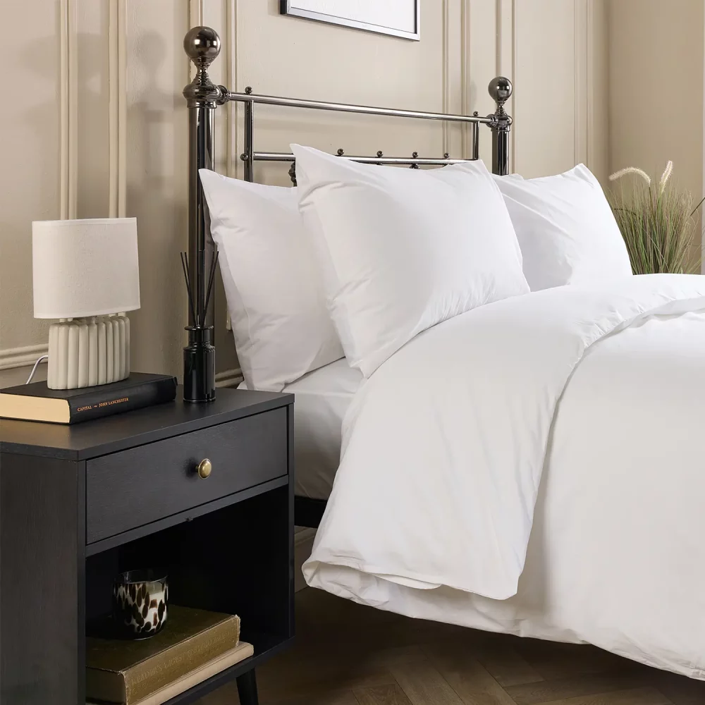 Hotel Pure Luxury cotton rich percale extra long duvet cover on a metal framed bed in a contemporary bedroom. There are also pillows on the bed. There is a dark wood cabinet with a lamp on top on the left-hand side of the room, with a painting above the bed.
