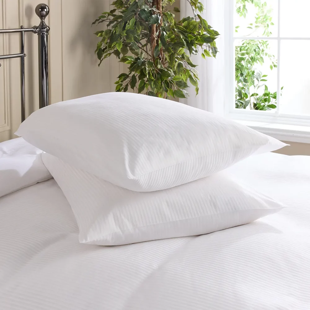 Hotel Pure Luxury cotton percale satin stripe pillowcases covering pillows that are sitting on a bed. There is a plant and window visible in the background.
