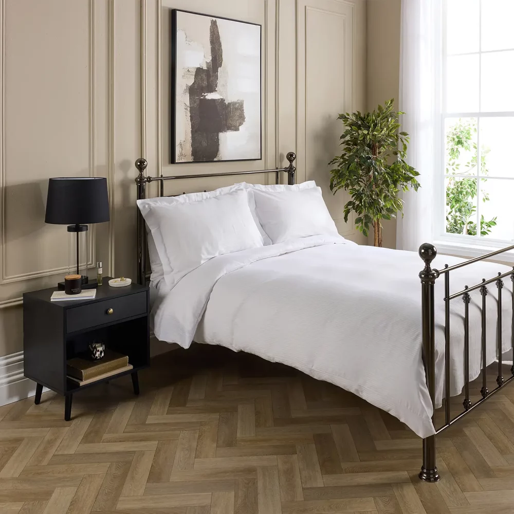A Hotel Pure Luxury cotton rich percale satin stripe duvet cover on a bed, complete pillows and pillowcases. There is a dark wood bedside cabinet with a lamp sitting on top of it next to the bed. You can see a contemporary painting above the metal framed bed.
