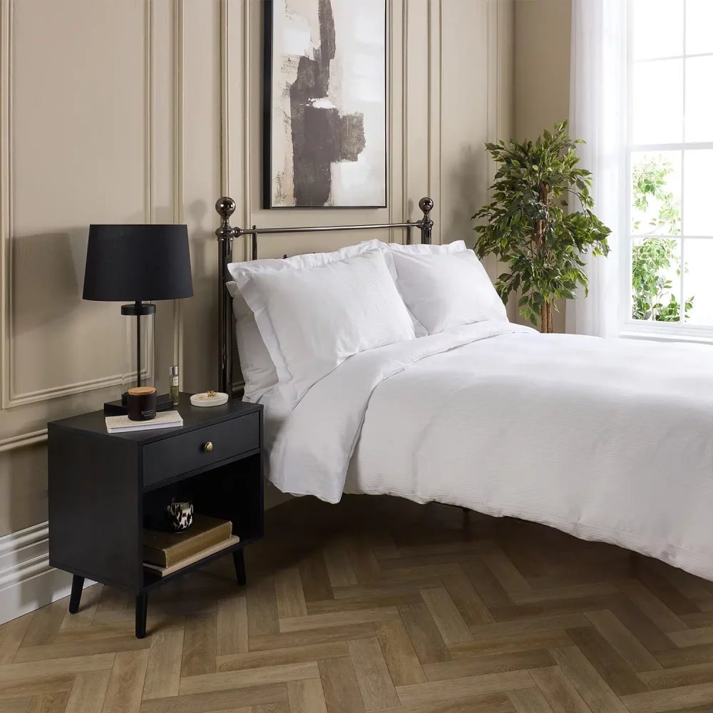 A Hotel Pure Luxury cotton rich percale satin stripe duvet cover on a bed, complete pillows and pillowcases. There is a dark wood bedside cabinet with a lamp sitting on top of it next to the bed. You can see a contemporary painting above the metal framed bed.