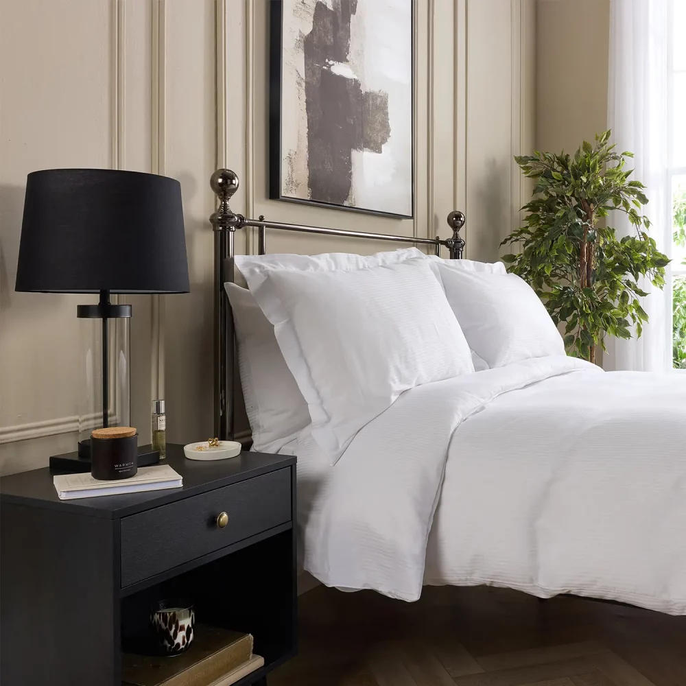 A Hotel Pure Luxury cotton rich percale satin stripe duvet cover on a bed, complete pillows and pillowcases. There is a dark wood bedside cabinet with a lamp sitting on top of it next to the bed. You can see a contemporary painting above the metal framed bed.
