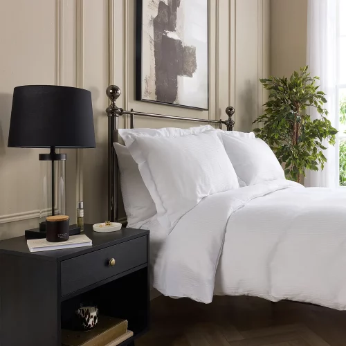 A Hotel Pure Luxury cotton rich percale satin stripe duvet cover on a bed, complete pillows and pillowcases. There is a dark wood bedside cabinet with a lamp sitting on top of it next to the bed. You can see a contemporary painting above the metal framed bed.
