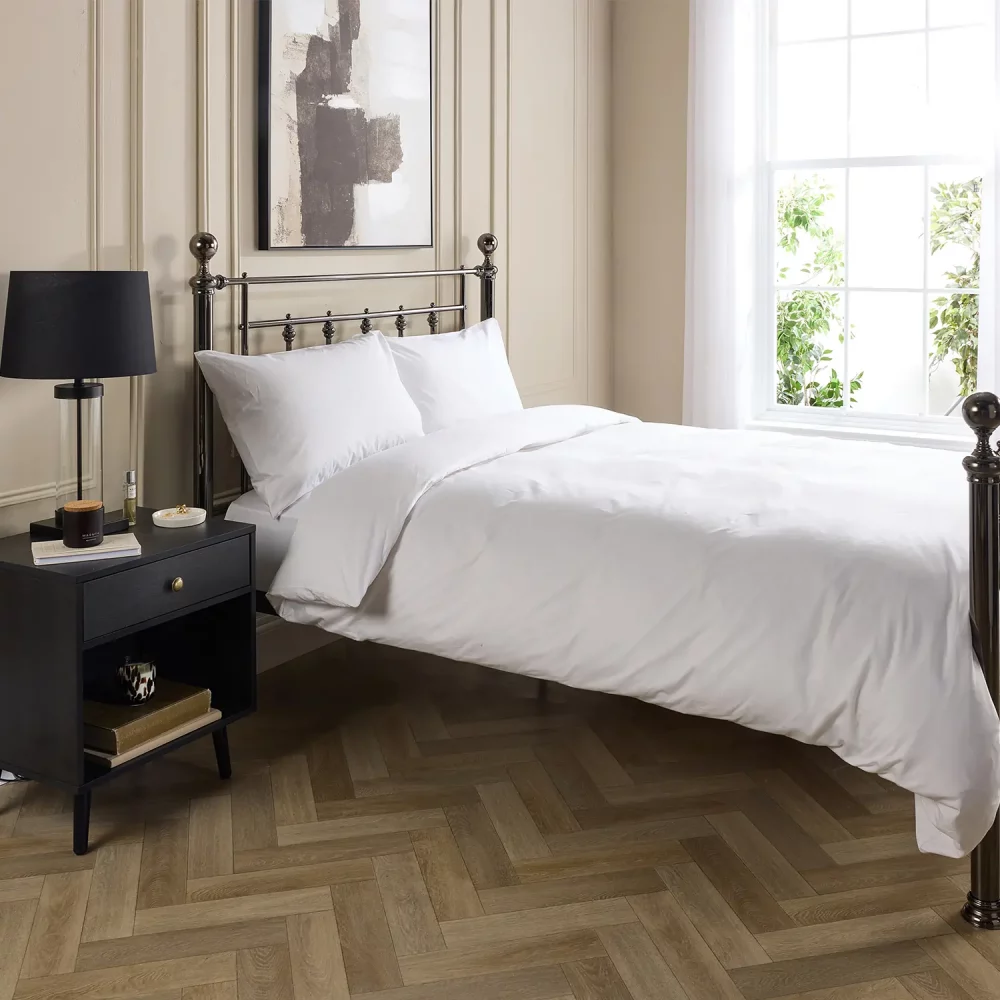 Hotel Pure Luxury cotton percale duvet cover. It's on a metal framed bed complete with white pillowcases. There is a dark wood bedside cabinet next to the bed with a lamp on top. A window is visible in the background and the bottom of a painting hanging above the bed is visible.