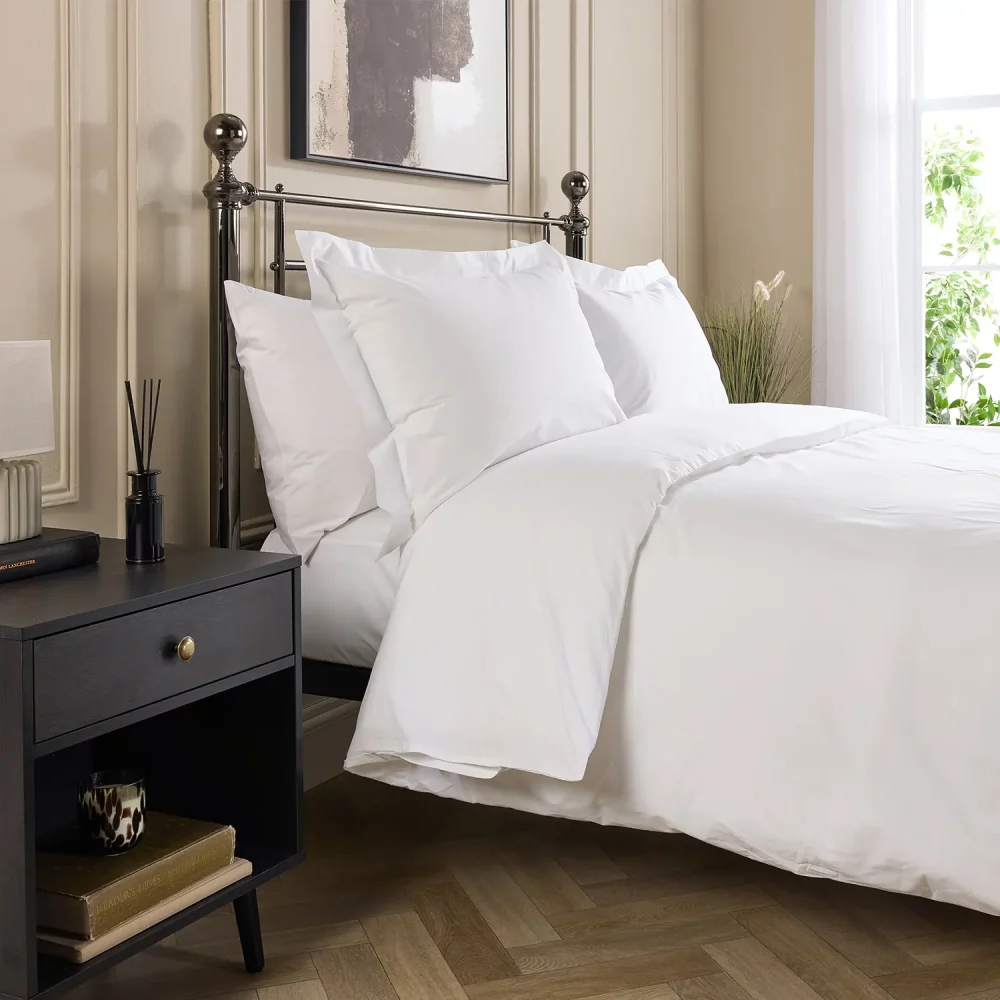 Hotel Pure Luxury cotton percale duvet cover. It's on a metal framed bed complete with white pillowcases. There is a dark wood bedside cabinet next to the bed with a lamp on top. A window is visible in the background and the bottom of a painting hanging above the bed is just about visible.