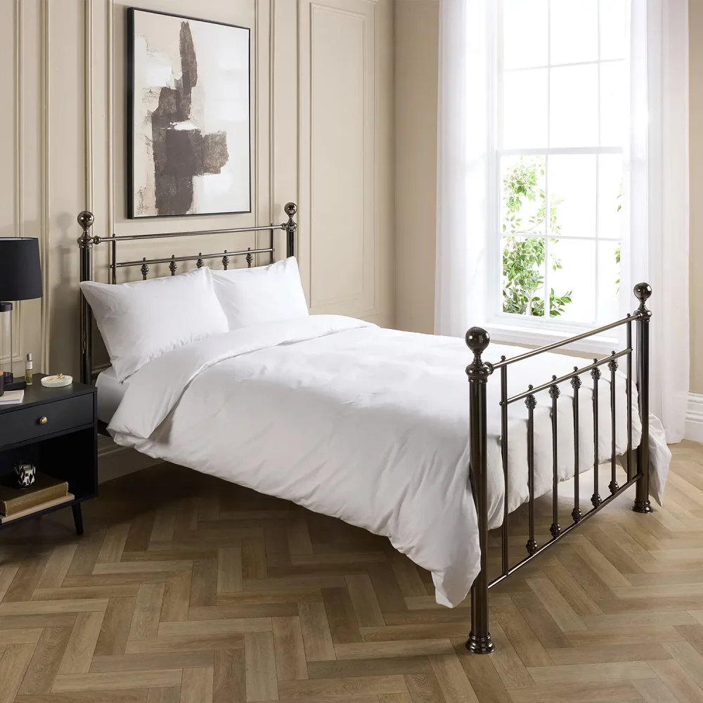 Hotel Pure Luxury cotton percale duvet cover. It's on a metal framed bed complete with white pillowcases. There is a dark wood bedside cabinet next to the bed with a lamp on top. A window is visible in the background and the bottom of a painting hanging above the bed is visible.