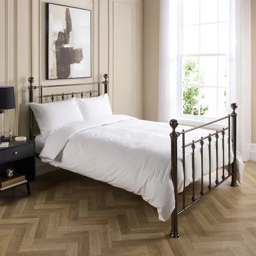 Hotel Pure Luxury cotton percale duvet cover. It's on a metal framed bed complete with white pillowcases. There is a dark wood bedside cabinet next to the bed with a lamp on top. A window is visible in the background and the bottom of a painting hanging above the bed is visible.